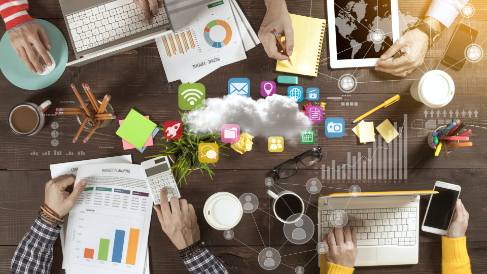 Top down view of table with laptops, tablets and reports with various business icons in the center