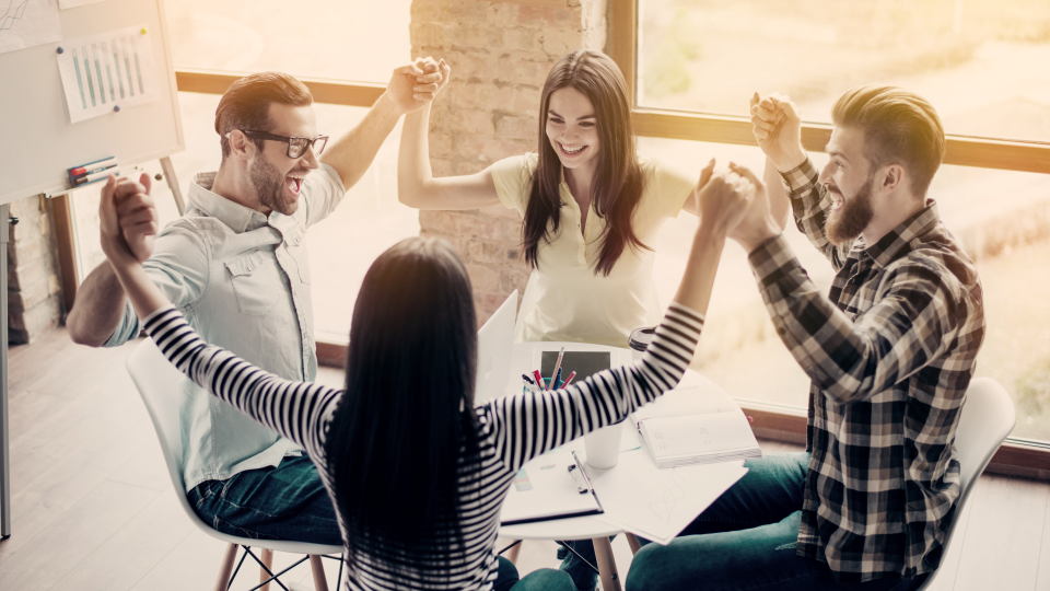 Four people sitting around a table with their arm raised in celebration holding hands