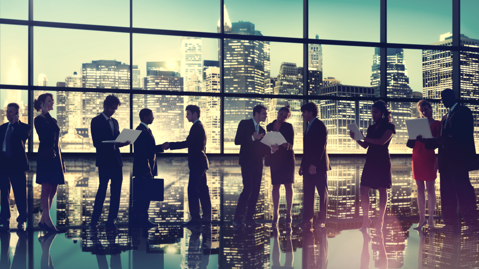 Many business people looking a papers and computers standing infornt of large window with cityscape in the background  